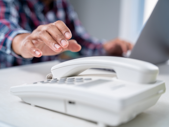 A person reaching for the phone on a desk with a laptop in the background